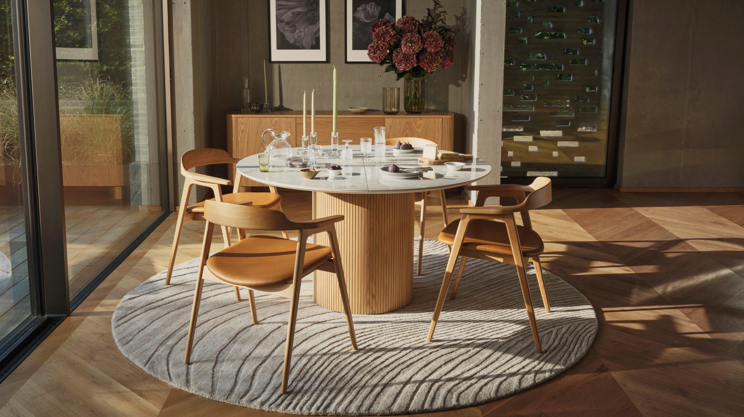 Moderní jídelní kout s kulatým dřevěným stolem Santiago, čtyřmi koženými židlemi Seoul, texturovaným kobercem a příborníkem s květinami a zarámovanými uměleckými tisky.